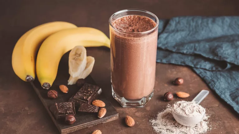 Shake proteico de chocolate ao lado de bananas, amêndoas, aveia e chocolate em pó